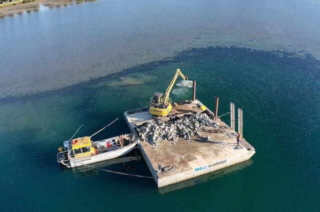 Aerial view of a barge with a yellow excavator on it and a boat tied to it. 