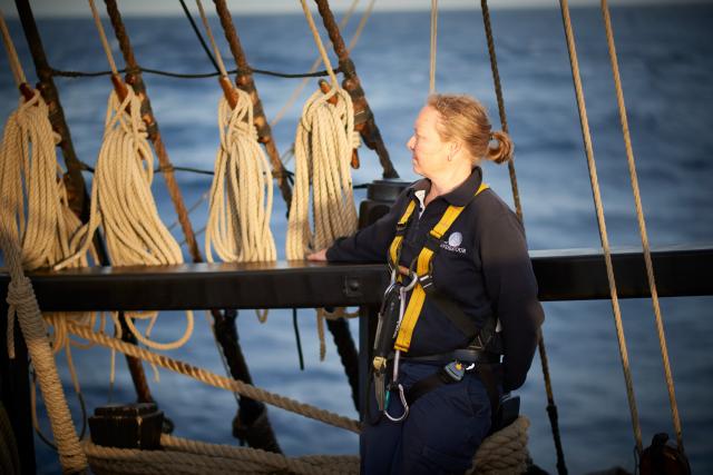 Women wearing a harness standing on a ship, with ropes and the ocean in the background
