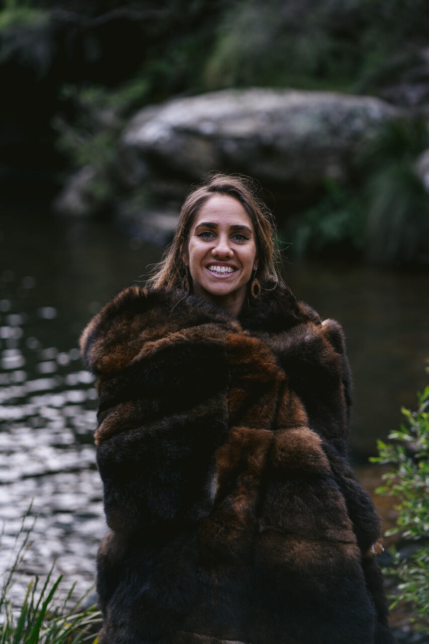 The poet Kirli Saunders is photographed by a river, wearing a beautiful possum skin cloak
