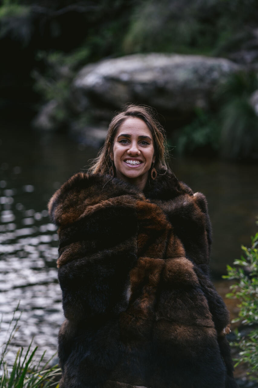 The poet Kirli Saunders is photographed by a river, wearing a beautiful possum skin cloak 