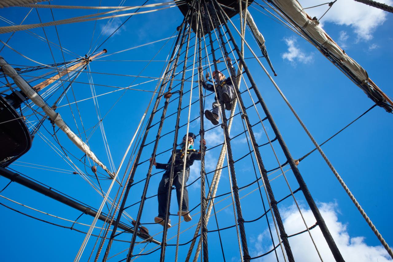 2 people climbing up the ships rigging (rope ladder) with a blue sky in the background 